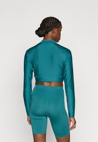 Σετ αθλητικών ρούχων σε τιρκουάζ χρώμα με κοντό τοπ μακρυμάνικο και ποδηλατικά σορτς με ψηλή μέση, κατασκευασμένα από λείο, ελαστικό ύφασμα με εφαρμοστή σχεδίαση.