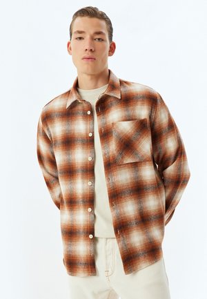 Camisa de franela en patrón de cuadros naranja, marrón y crema; cuenta con un bolsillo, cierre de botones y un ajuste relajado. Lleva una camiseta beige debajo.