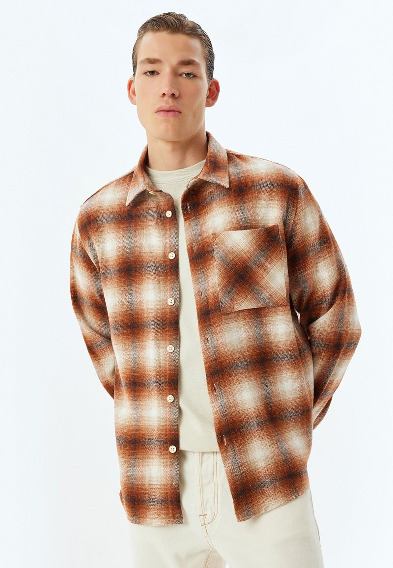 Flanellen shirt in oranje, bruin en crèmekleurig ruitpatroon; beschikt over een zak, knopenfront en een relaxte pasvorm. Draagt over een beige top.