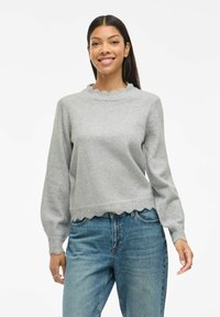 Leende kvinna i en ljusgrå stickad tröja med vågiga kanter och blå denimjeans, som står mot en vit bakgrund.