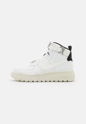 Baskets montantes blanches avec semelle écrue, col rembourré noir, laçage avant, logo swoosh latéral et bride de cheville avec boucle en métal.