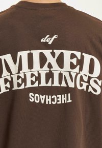 Brun T-skjorte med store hvite trykte bokstaver som leser "def MIXED FEELINGS" på baksiden. Myk, bomullsblanding med en avslappet passform.