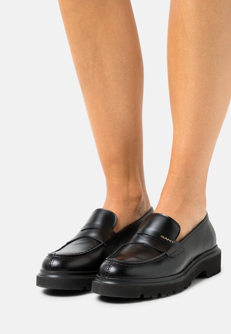 Mocassins noirs en cuir à enfiler avec semelles épaisses, portés pieds nus sur un fond clair uni.