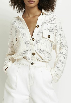 Nainen, jolla on valkoinen pitsinen nappipaita, jossa on kaksi rintataskua ja valkoiset korkeavyötäröiset housut, kädet taskuissa.