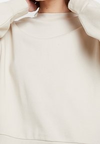 Felpa oversize color crema con collo alto, polsini a costine e texture del tessuto liscia. Design semplice e minimalista.