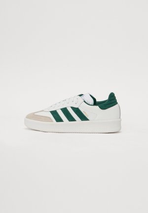 Weißer Sneaker mit niedrigem Schaft, grünen Wildlederstreifen, beiger Zehenkappe, gepolstertem Schaft und weißer Sohle, seitlich auf weißem Hintergrund betrachtet.