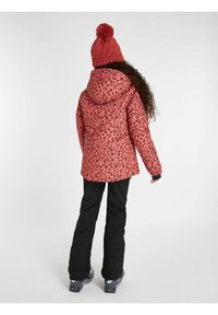 Doudoune rose à motif avec capuche et pompon en tricot, associée à un pantalon de ski noir. Accent sur la chaleur et la fonctionnalité en extérieur.