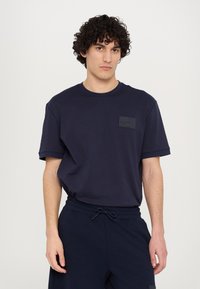 Marineblå bomulds T-shirt med korte ærmer, rund hals og en struktureret sort lap på venstre bryst. Casual pasform og glat tekstur.