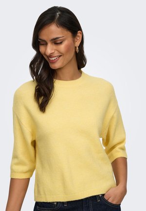 Femme aux cheveux foncés portant des boucles d'oreilles créoles dorées et un pull à manches courtes jaune clair, souriante et regardant vers le bas, une main dans la poche.
