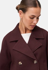 Femme portant un manteau brun foncé croisé avec de grands revers et des boucles d'oreilles en or, regardant vers le bas sur son côté gauche.