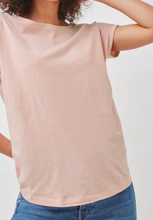 Femme portant un t-shirt rose clair à manches courtes et un jean bleu, un bras levé et des cheveux bouclés partiellement visibles.