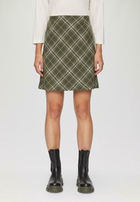 Spódnica w kratę w oliwkowym kolorze o rozkloszowanym kształcie, z białymi i ciemnozielonymi liniami, zestawiona z czarnymi botkami za kostkę. Gładka tekstura.