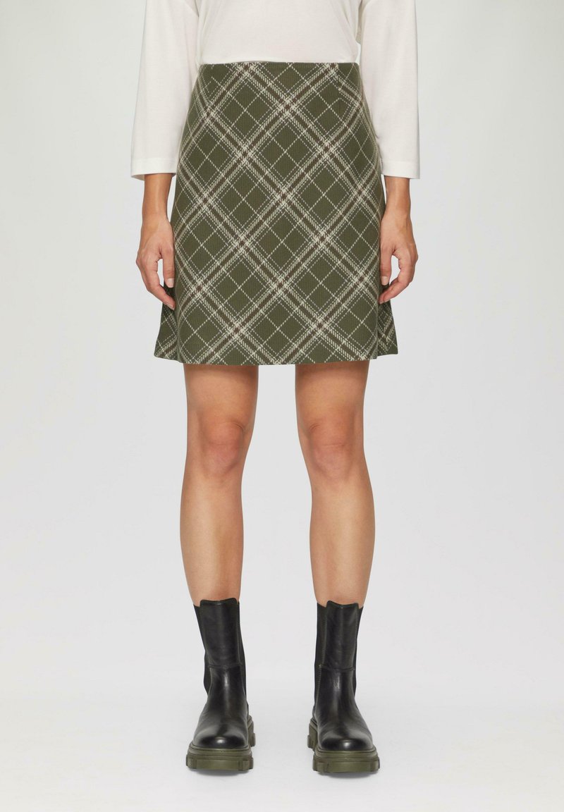 Spódnica w kratę w oliwkowym kolorze o rozkloszowanym kształcie, z białymi i ciemnozielonymi liniami, zestawiona z czarnymi botkami za kostkę. Gładka tekstura.