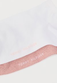 Λευκή βαμβακερή κάλτσα με ροζ, υφασμάτινο μανσέτα. Η λέξη "TOMMY HILFIGER" είναι τυπωμένη με αντίθετο λευκό χρώμα πάνω τόσο στο λευκό όσο και στο ροζ μέρος.