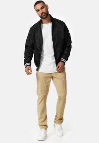 Chaqueta bomber negra con puños a rayas, camiseta blanca, pantalones caquis y zapatillas blancas, de pie sobre un fondo neutro.