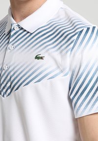 Vit polo-skjorta med blå gradientränder, korta ärmar och klassisk krage. Har en grön krokodil-logotyp på bröstet.