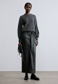 Hall triiksõrmikuga kampsun, kõrge vöökohaga oliivirohelisest nahast maxi seelik paelaga ja mustad kingad; hoiab käes musta käekotti.