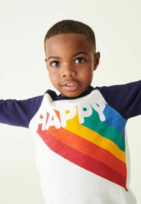 Fehér és tengerészkék raglán póló, nagy "HAPPY" felirattal, vastag betűkkel, és átlós szivárványcsíkokkal piros, narancs, sárga, zöld és kék színekben.