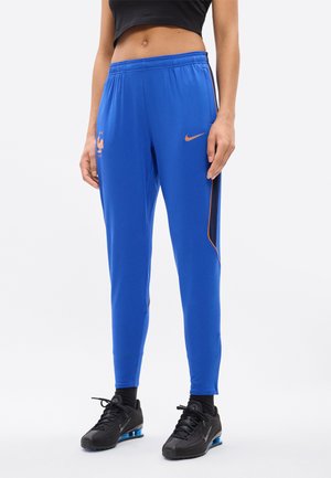 Personne portant un pantalon de sport Nike bleu avec des logos orange et des baskets noires, debout devant un fond clair uni.