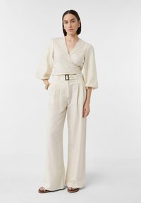Jasno beżowa bluzka o kroju kopertowym z bufkami, zestawiona z spodniami z szerokimi nogawkami o wysokim stanie. Gładki materiał, prosta pasek i brązowe sandały na obcasie.