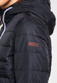 Černá péřová bunda s texturovaným povrchem, vybavená kapucí a bílým stahovacím šňůrkem. Na rukávu je hnědá kožená logo nášivka.