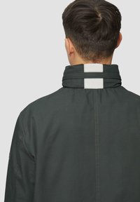 Dunkelgrüne Jacke mit hohem Kragen und einem weißen Akzentstreifen. Der Stoff wirkt glatt, mit sichtbaren Nähten an den Säumen.