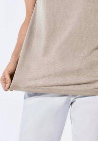 Beige mouwloos topje van een zachte, gestructureerde stof, vastgehouden door een hand. Witte broek gedeeltelijk zichtbaar aan de onderkant, wat een lichte kleurcontraster creëert.