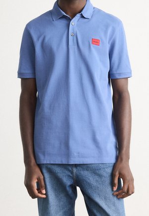 Man wearing a light blue short-sleeve polo shirt with a red "Hugo" logo patch and blue jeans, standing against a plain background.