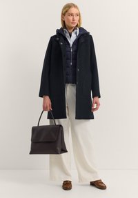 Dunkler Wollmantel mit Kapuze, ausgestattet mit Knöpfen und Taschen, über einer navyblauen Steppjacke getragen. Kombiniert mit cremfarbenen Hosen und einer dunklen Handtasche.