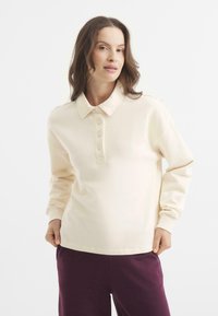 Kremowy sweter z kołnierzykiem, pięcioma guzikami, długimi rękawami i ściągaczami na mankietach. Wykonany z miękkiego materiału, o luźnym kroju.