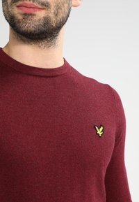 Bordeauxfarbener Strickpullover mit rundem Ausschnitt und subtiler Textur, ausgestattet mit einem kleinen gelben gestickten Logo auf der linken Brust.