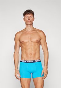 Blå boxer-shorts lavet af blødt stof med en marineblå talje, der har "PUMA" logoet trykt i hvid. Modellens forside vises.