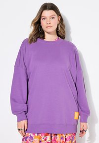 Sweatshirt violet oversize avec des manches longues, un col rond et un patch logo jaune sur le côté. Tissu doux avec une texture lisse.