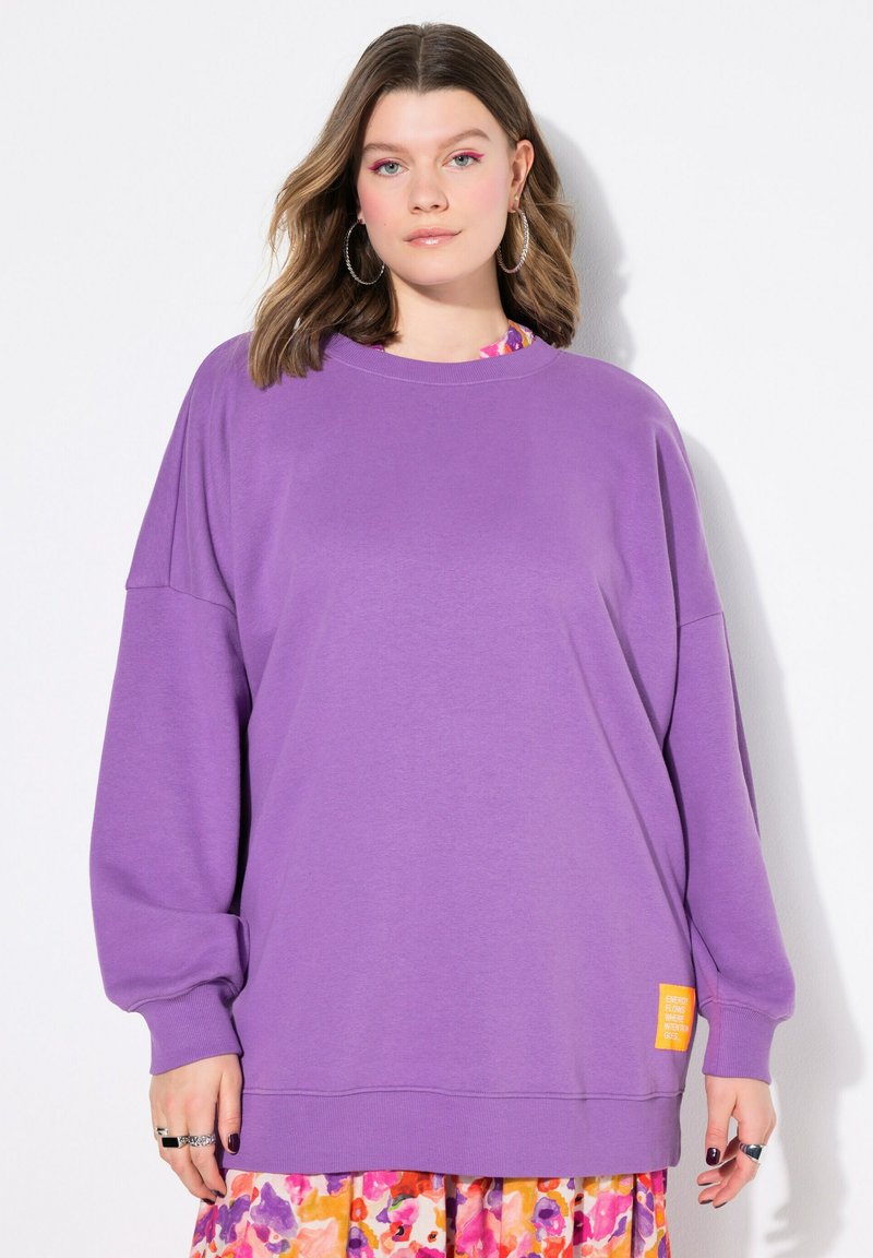 Sweatshirt violet oversize avec des manches longues, un col rond et un patch logo jaune sur le côté. Tissu doux avec une texture lisse.