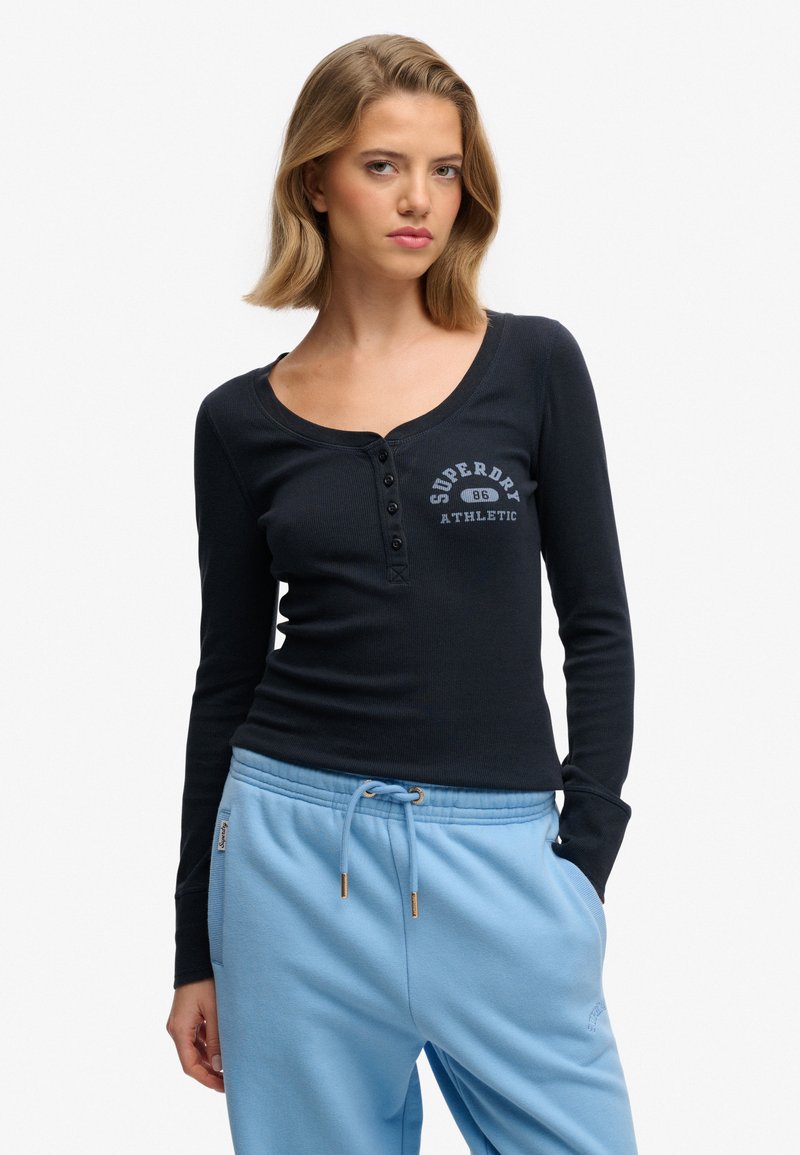 T-shirt noir à manches longues avec un col arrondi, des détails à boutons et un graphisme blanc "Superdry Athletic" sur la poitrine, assorti à un pantalon de jogging bleu.