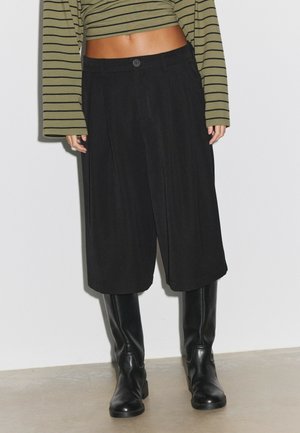 Persona che indossa pantaloni neri a gamba larga e tagliati, stivali neri al ginocchio e una maglia verde corta a maniche lunghe con righe orizzontali nere.