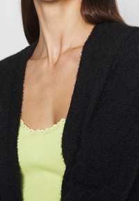 Svart stickad kofta med mjuk textur, med en djup V-ringning och öppen framdel, över en ljusgrön ribbad linne med spetsdetalj.