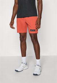 Orange Badehosen mit einem elastischen Bund und seitlichen Taschen; schwarzes Logo-Patch am linken Bein; kombiniert mit weißen Sneakers und Socken.