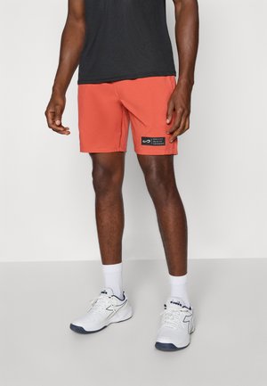 Orange swim shorts with an elastic waistband and side pockets; black logo patch on the left leg; paired with white sneakers and socks.