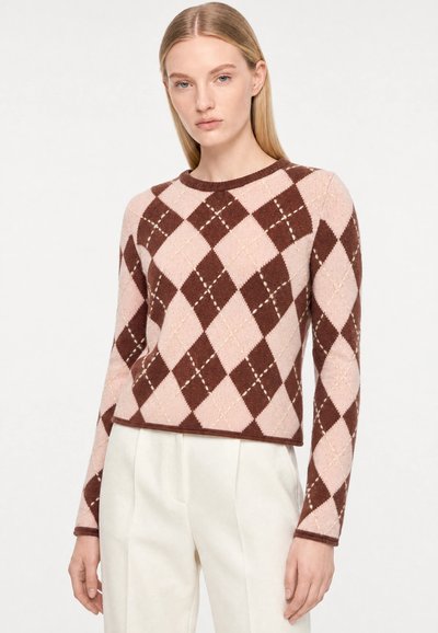 Femme portant un pull à manches longues à motif losanges roses et marron avec un pantalon blanc taille haute, regardant vers l’avant.