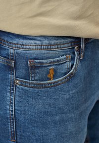 Vaqueros de mezclilla azul con una superficie texturizada, que presentan un pequeño logo marrón en el bolsillo trasero y costuras naranjas contrastantes a lo largo de las costuras.