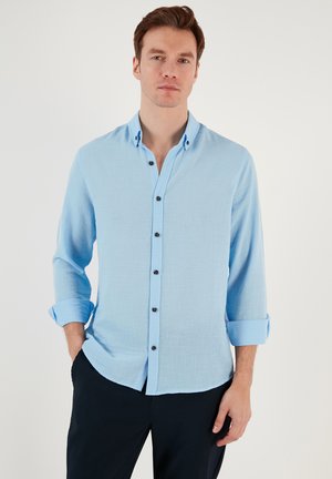 Camisa azul claro de botones hecha de una tela texturizada, con un cuello clásico, mangas largas arremangadas en los puños y botones oscuros.