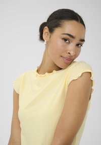 T-shirt jaune à manches courtes avec un col festonné. Le tissu est lisse avec une finition légère et texturée.