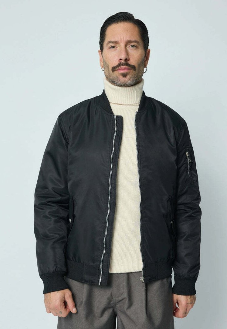 Veste bomber noire en tissu lisse avec une fermeture éclair à l'avant, des poignets côtelés et une poche zippée sur la manche gauche. Portée sur un col roulé crème.