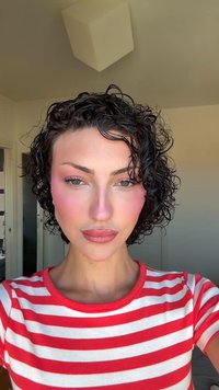 Des cheveux bouclés et sombres encadrent un visage aux pommettes bien définies. Vêtue d'un t-shirt rayé rouge et blanc, ses lèvres sont d'un rose mat et ses joues légèrement rosies.
