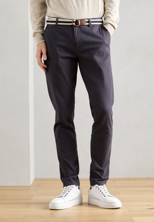 Marineblauwe katoenbroek met een slim fit, voorzien van een gestreepte riem en een bruine gesp, gecombineerd met witte sneakers op een houten vloer.