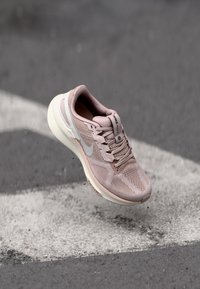 Light pink athletic shoe with a mesh upper, rounded shape, grey accents, and a white sole, shown floating above a gray background.