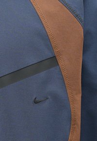 Nike jack met een blauw en bruine kleurstelling, gemaakt van textuurstof, met contrasterende stiksels en een zwarte horizontale accentband.