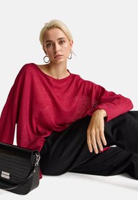 Přehnaně velký, průhledný červený pletený svetr s širokým výstřihem a dlouhými rukávy, spárovaný s volnými černými kalhotami a černou kabelkou s krokodýlím vzorem.