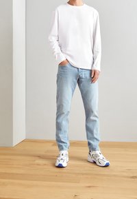 Camisa blanca de manga larga, jeans de color azul claro y zapatillas blancas con acentos en azul y negro. Conjunto simple y casual sobre un suelo de madera.